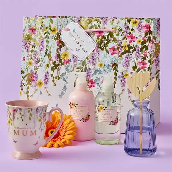 A floral Mother’s Day gift set featuring a decorative mug, pastel bath products, and a blue reed diffuser, arranged in front of a flower‑covered gift bag with a festive tag.